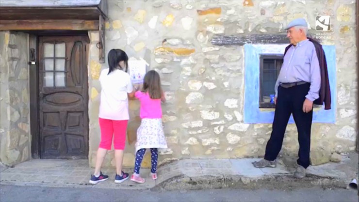 Dos niñas cuelgan un cartel en la fachada de una casa en presencia de un señor mayor.